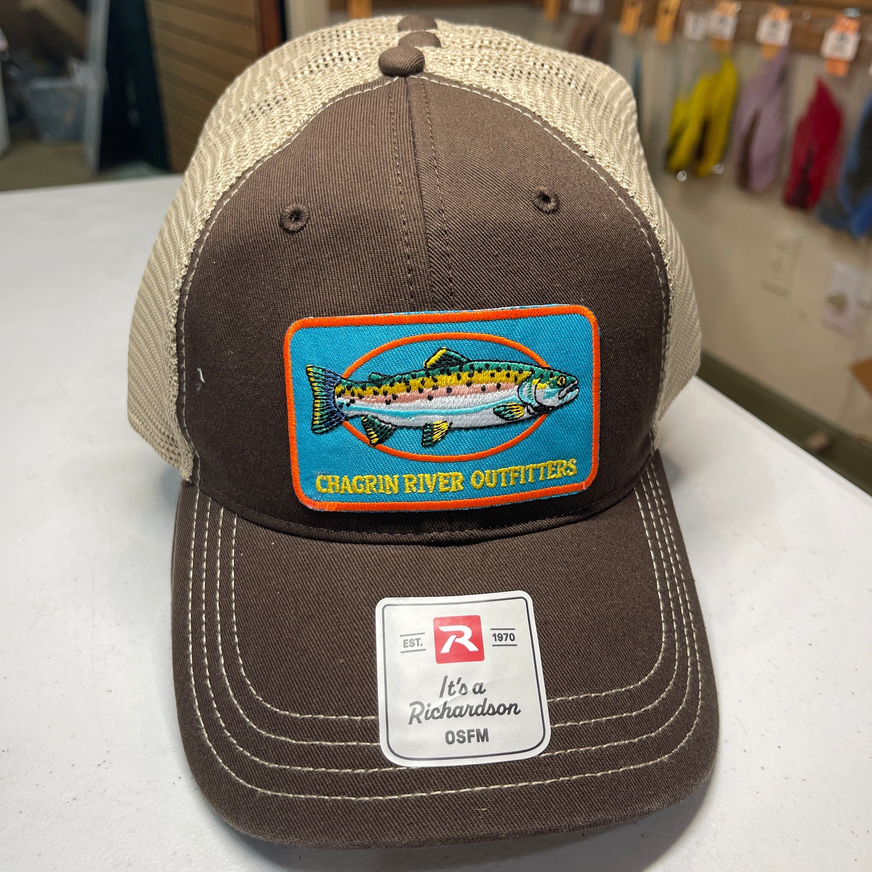 A brown and beige trucker hat with a colorful fish patch reading 'Chagrin River Outfitters' and a visible Richardson CSPM sticker on the brim, photographed on a tabletop in a store.