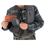 Person wearing a gray Simms Freestone Vest holding an orange and gray pouch with a brand logo.