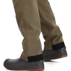 Brown Simms MEN's Rogue Softshell Pants with black cuffs and gray rubber boots on a white background