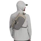 Person wearing a beige and gray Simms Headwaters Sling Pack bag on a white background