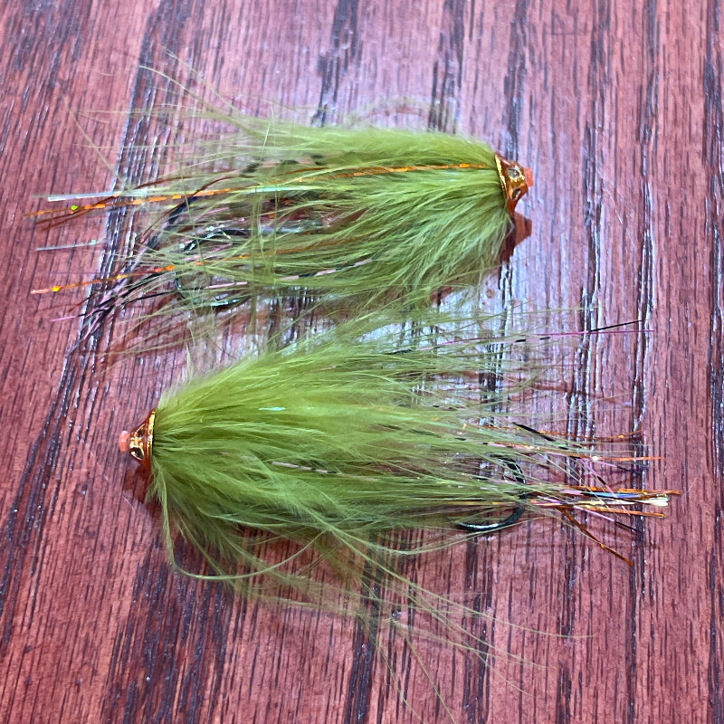 Two green fly fishing lures with copper-colored hooks and wispy hair strands, displayed on a dark brown wood-grain surface.