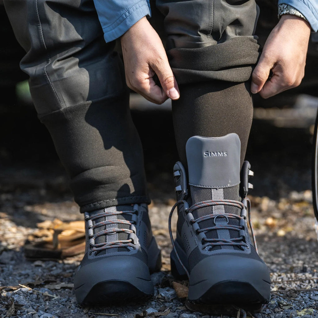 Person wearing black hiking boots with a visible brand logo on a natural ground surface.