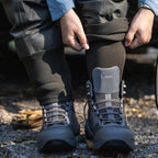 Person wearing black hiking boots with a visible brand logo on a natural ground surface.