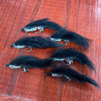 A collection of artificial fishing flies with white, black, and olive colors distributed across the frame, displayed on a wooden surface.