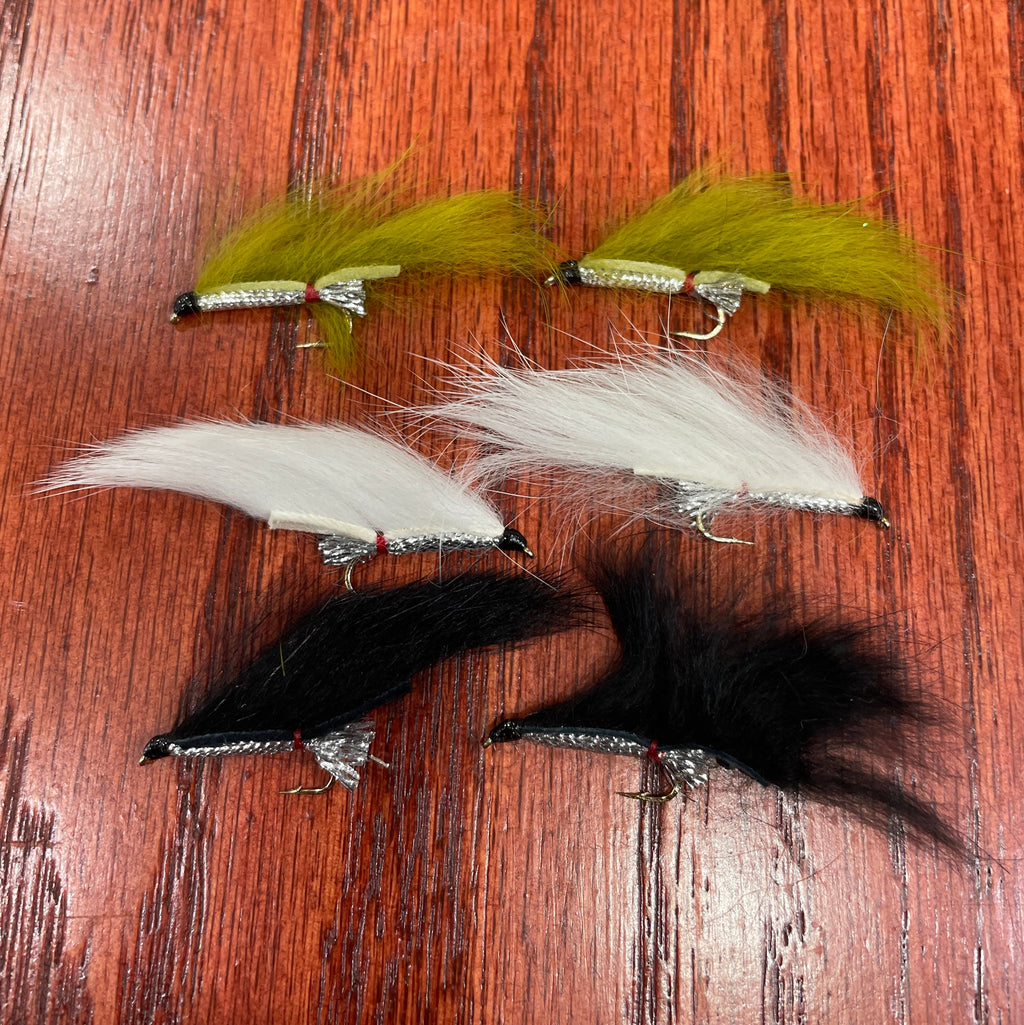 A collection of artificial fishing flies with white, black, and olive colors distributed across the frame, displayed on a wooden surface.
