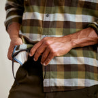Person wearing a plaid Orvis MEN's Flat Creek Tech Flannel Shirt holding a black bag against a neutral background