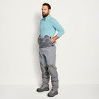 An angler wearing a pair of Orvis men's PRO LT fishing waders in gray color with adjustable elastic suspenders, neoprene booties, and a stretchy external pocket.