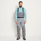 An angler wearing a pair of Orvis men's PRO LT fishing waders in gray color with adjustable elastic suspenders, neoprene booties, and a stretchy external pocket.