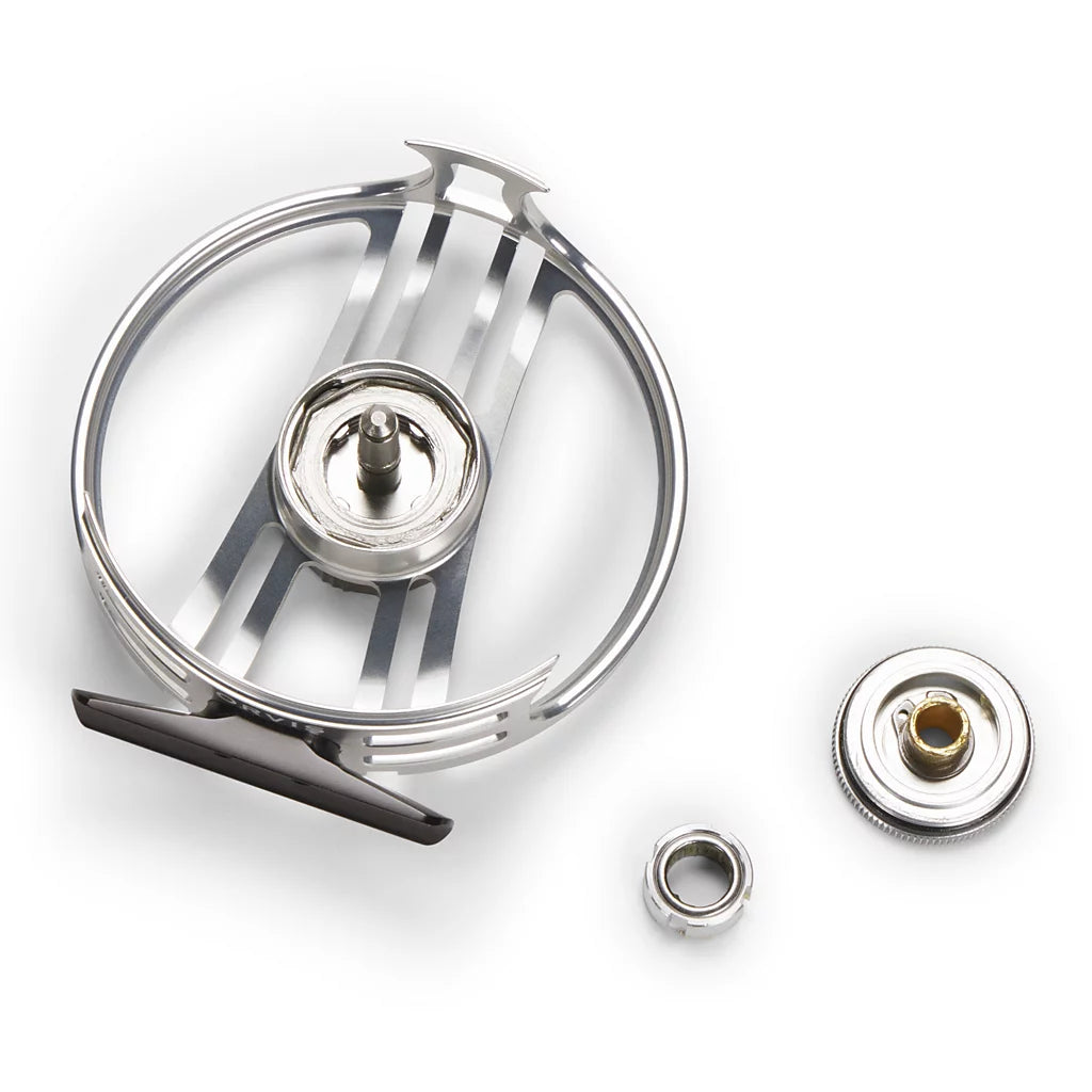 A stainless steel fly tying or fishing component set on a white background, featuring a circular metal frame, a central spool, a small ring, and a separate circular piece with a brass-colored center.
