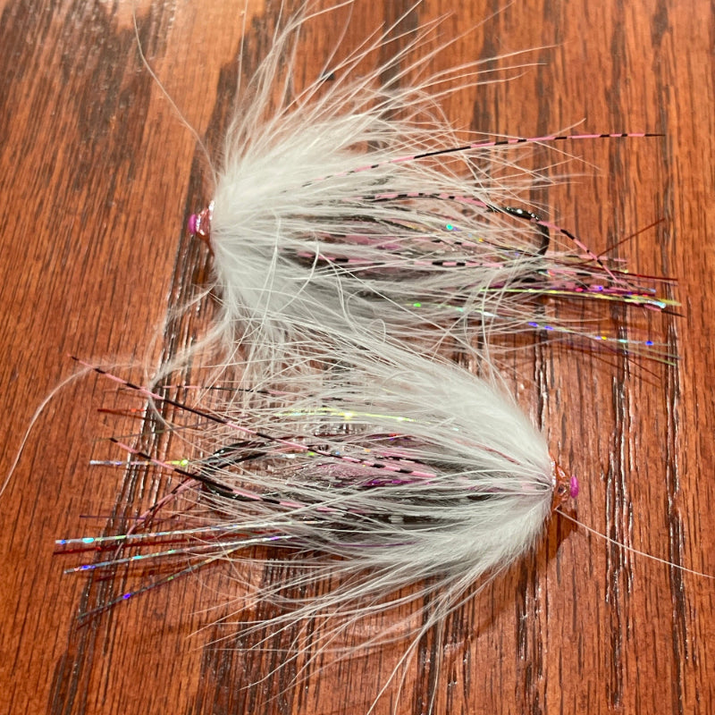 Two handmade tube fly fishing lures made from white hackle feathers and pink thread, resting on a brown wooden surface.