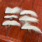 A collection of artificial fishing flies with white, black, and olive colors distributed across the frame, displayed on a wooden surface.