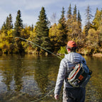 Person fly fishing in a serene forest lake setting wearing a Green and orange Fishpond Stormshadow Sling Pack
