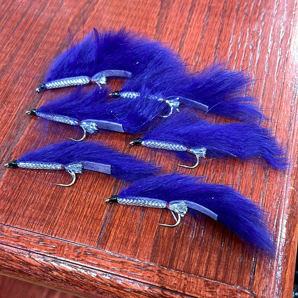 A collection of artificial fishing flies with white, black, and olive colors distributed across the frame, displayed on a wooden surface.