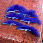 A collection of artificial fishing flies with white, black, and olive colors distributed across the frame, displayed on a wooden surface.