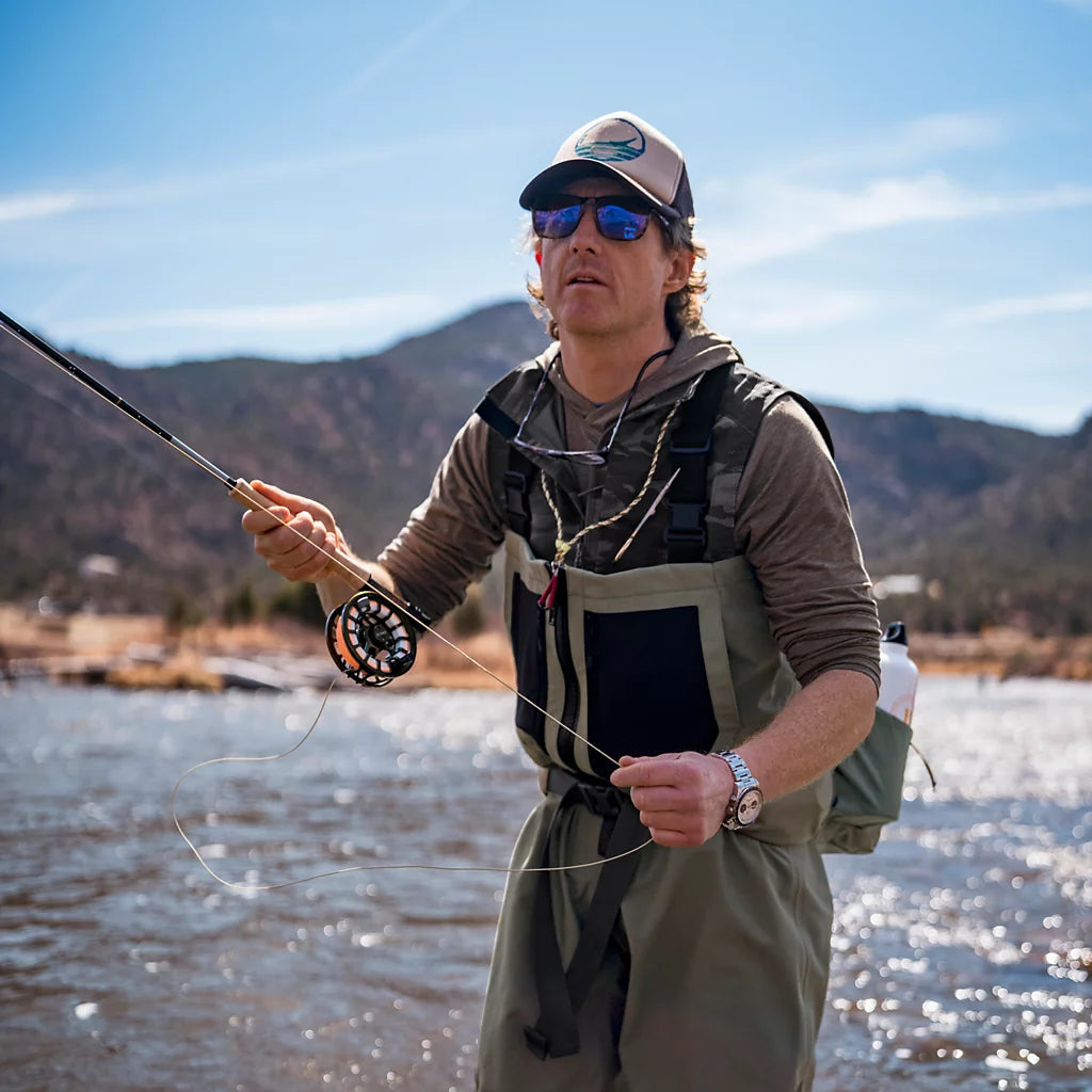A man wearing Orvis Clearwater Zip Waders, a matching cap, sunglasses, and a harness stands in a river holding a fly rod, with mountains and blue sky in the background.