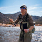 A man wearing Orvis Clearwater Zip Waders, a matching cap, sunglasses, and a harness stands in a river holding a fly rod, with mountains and blue sky in the background.