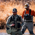 Two men in waders and fly-fishing gear standing in shallow water with dry reeds in the background. The man in front wears a dark camo vest over a gray long-sleeve top and a dark cap, while the man behind wears an orange shirt and gray waders.