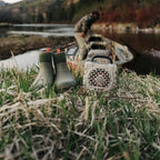 Green rubber boots and a Turtlebox Cub speaker on grass near a body of water.