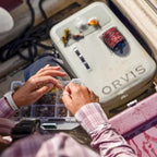 Person organizing fishing lures in a tackle box next to an Orvis Pro boat bag.