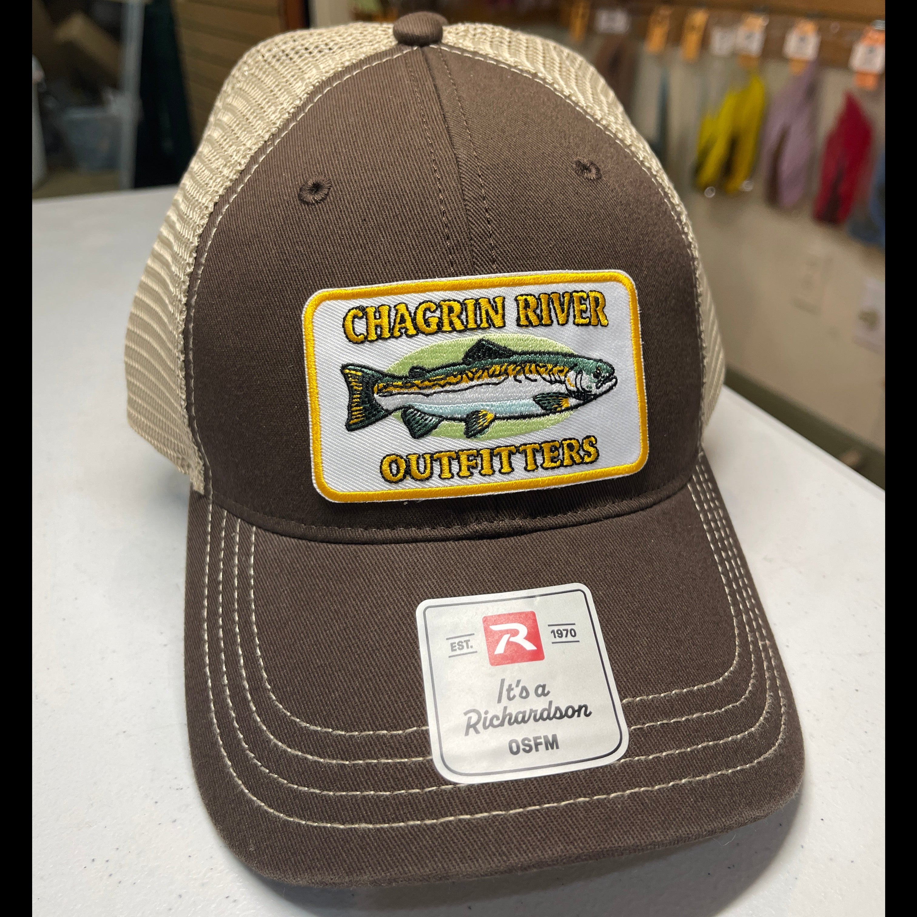 A brown and beige trucker hat with a 'Chagrin River Outfitters' fish patch on the front, resting on a white surface with a store background.