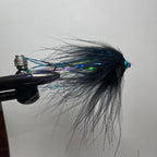 A close-up of a tube fishing fly tied with black hackle feathers and iridescent blue, green, and purple fibers, held in a vise against a plain light background.