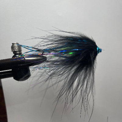 A close-up of a tube fishing fly tied with black hackle feathers and iridescent blue, green, and purple fibers, held in a vise against a plain light background.