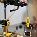 An ergonomic Loon bobbin with a yellow and black powder-coated handle, a metal spool holder, and a long metal shaft together with a fly tying vise in the background. 