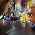 An ergonomic Loon bobbin with a yellow and black powder-coated handle, a metal spool holder, and a long metal shaft and a fly tying vise in the background. 