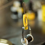 An ergonomic Loon bobbin with a yellow and black powder-coated handle, a metal spool holder, and a long metal shaft.