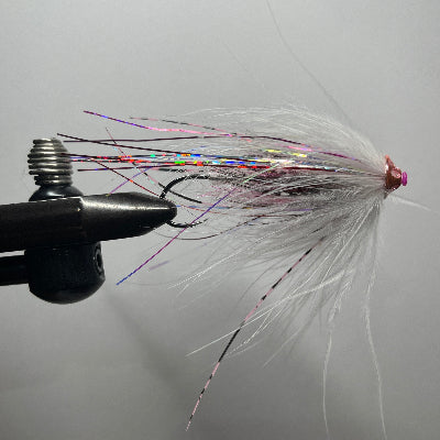 A close-up of a tube fishing fly tied in a black vise, featuring white feathers, holographic rainbow glitter, and a pink head, set against a plain gray background.