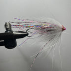 A close-up of a tube fishing fly tied in a black vise, featuring white feathers, holographic rainbow glitter, and a pink head, set against a plain gray background.