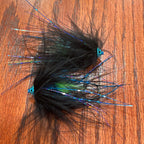 Two black fly fishing lures with iridescent blue, green, and purple hair wings, displayed on a brown wooden surface.