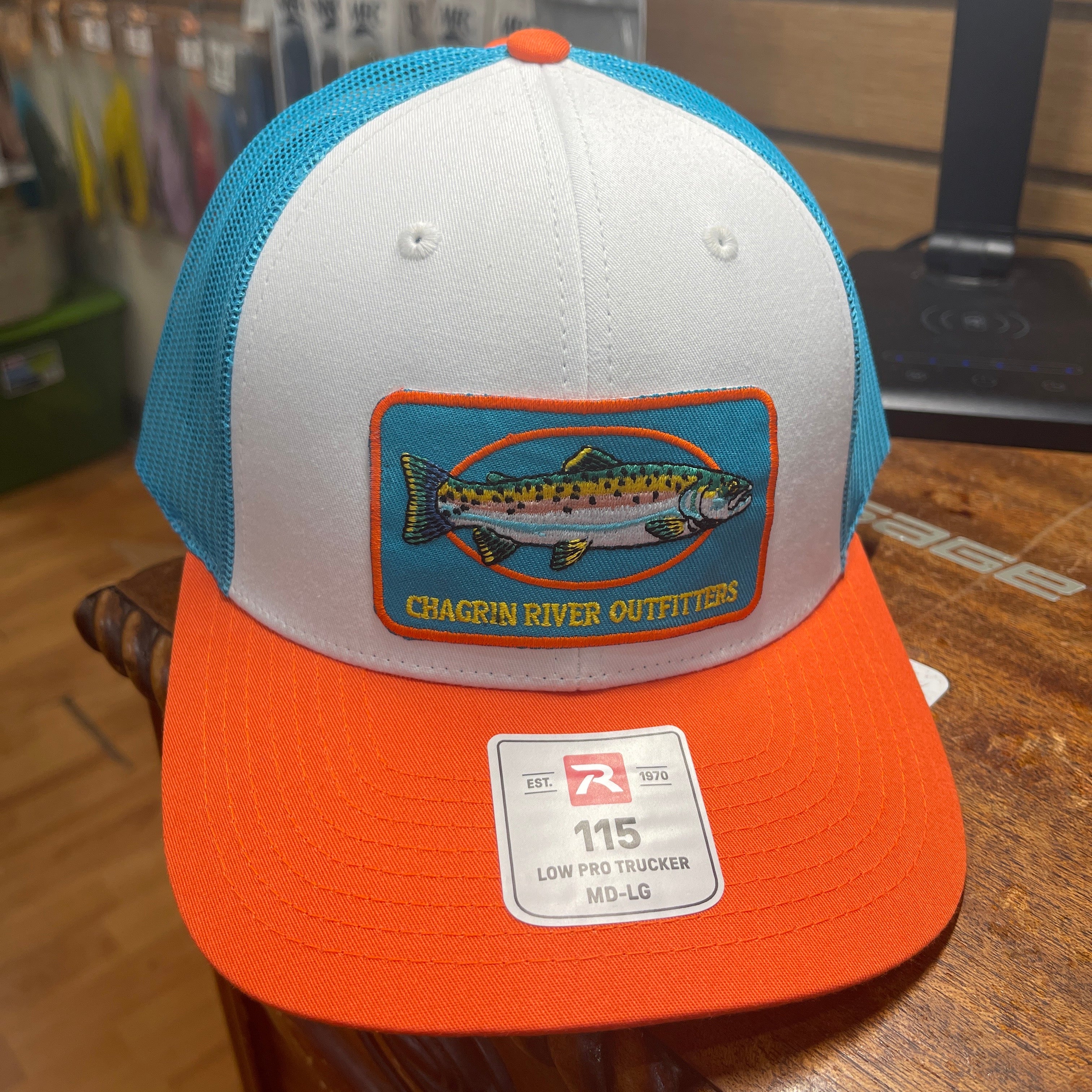 A low-profile trucker hat with a white front panel, turquoise mesh back, and bright orange brim. It features a rectangular embroidered patch of a chagrin river fish with the text 'CHAGRIN RIVER OUTFITTERS' and a white size sticker on the brim.