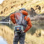 Fishing angler wearing a green Fishpond Thunderhead Small Submersible Lumbar pack with a brown shoulder strap and zippered closures, featuring a logo on the front and a net on the back.