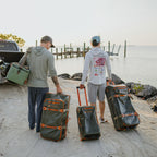 Two men with large Green Fishpond Stormshadow Medium Rolling Duffel with orange straps on a white background on a dock by a body of water.