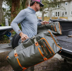 Man loading large green duffel bags with orange straps into a truck bed.