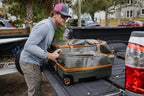 A man is loading a rolling Fishpond Stormshadow Rolling Carry-On with fishing gear into the bed of a pickup truck.