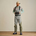 A man wearing Orvis breathable fly-fishing waders in gray and green colors, featuring external pockets and neoprene booties, standing against a plain background.
