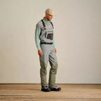 A man wearing Orvis breathable fly-fishing waders in gray and green colors, featuring external pockets and neoprene booties, standing against a plain background.