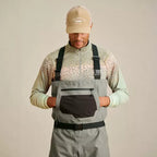 A man wearing Orvis breathable fly-fishing waders in gray and green colors, featuring external pockets and neoprene booties, standing against a plain background.
