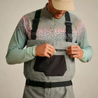 A man wearing Orvis breathable fly-fishing waders in gray and green colors, featuring external pockets and neoprene booties, standing against a plain background.