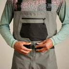 A man wearing Orvis breathable fly-fishing waders in gray and green colors, featuring external pockets and neoprene booties, standing against a plain background.