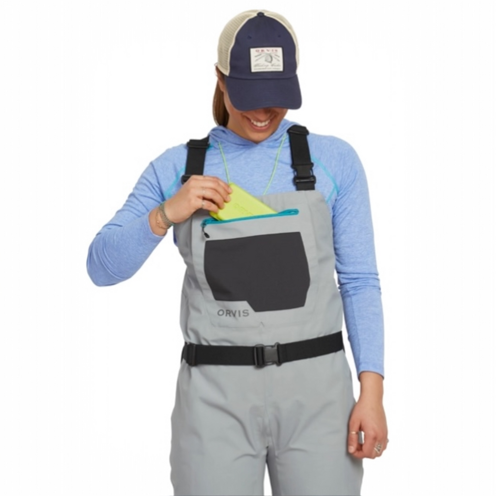 Person wearing gray Orvis women waders with suspenders on a white background while getting a fly box from the front pocket. 