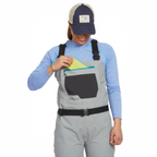Person wearing gray Orvis women waders with suspenders on a white background while getting a fly box from the front pocket. 