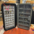 Open fly-fishing fly box with various dry flies and nymphs on a wooden surface