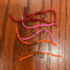 Six squirmy worms fly fishing flirs in red, pink, and orange on a wooden surface