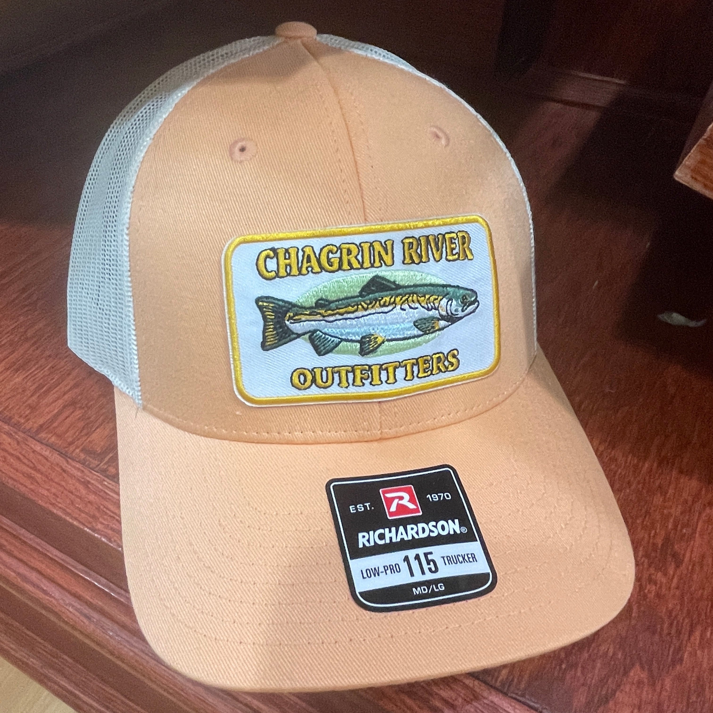 A tan and white trucker hat with a 'Chagrin River Outfitters' fish patch on the front and a 'Richardson 115' size label on the side, photographed on a dark wood floor.