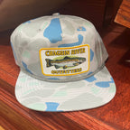 A camouflage baseball cap with a light gray, blue, and mint green pattern, featuring a front patch that reads 'CHAGRIN RIVER OUTFITTERS' with a fish illustration. The cap is resting on a polished wooden surface.