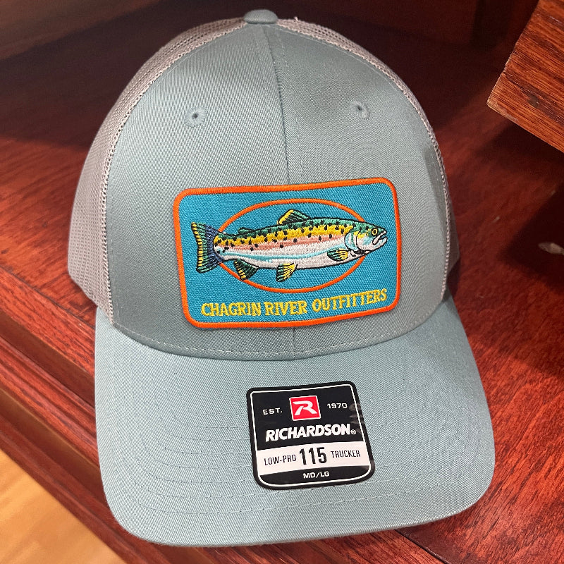 A light gray Richardson 115 trucker hat with a colorful fish patch on the front, resting on a dark wood surface.