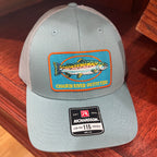 A light gray Richardson 115 trucker hat with a colorful fish patch on the front, resting on a dark wood surface.